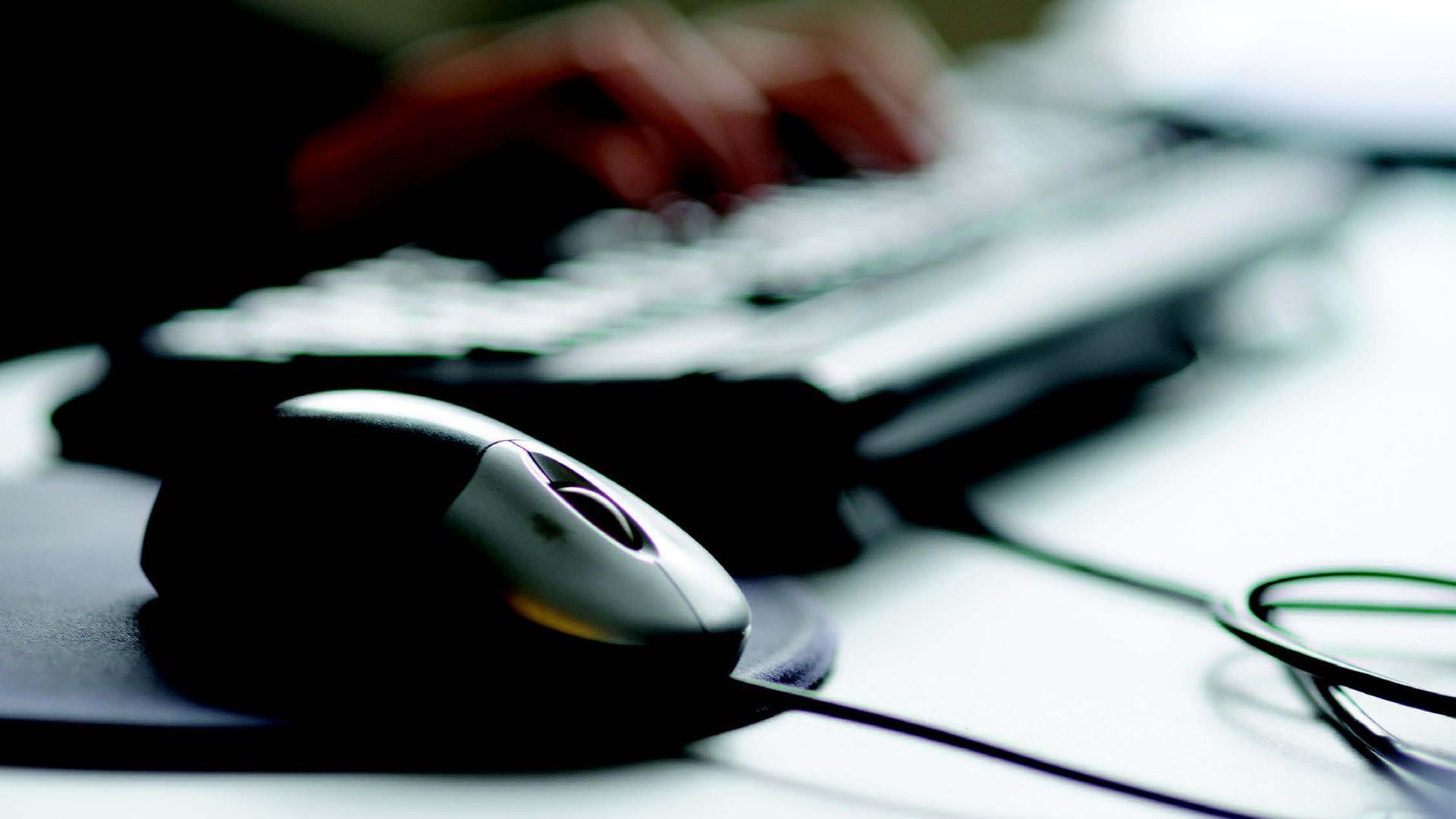 Computer mouse on pad with keyboard and hands typing in the background