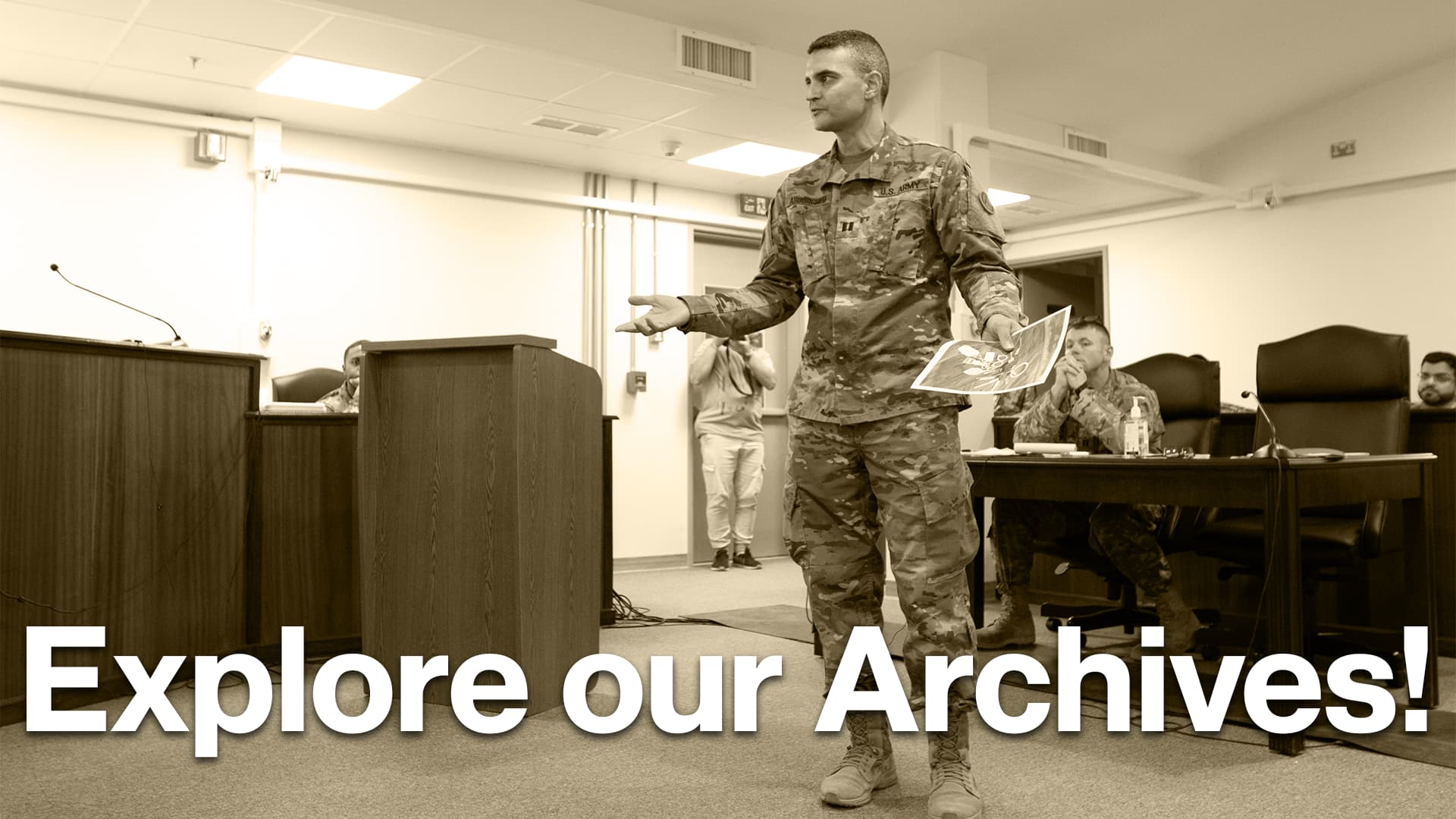 A U.S. Army officer in combat uniform stands in front of a podium, gesturing while holding two printed images. He is giving a presentation in a conference room. Behind him, another service member is seated at a table, observing the presentation, while a third person takes a photo in the background. The room is furnished with wooden desks and chairs, and the environment suggests a formal military meeting.