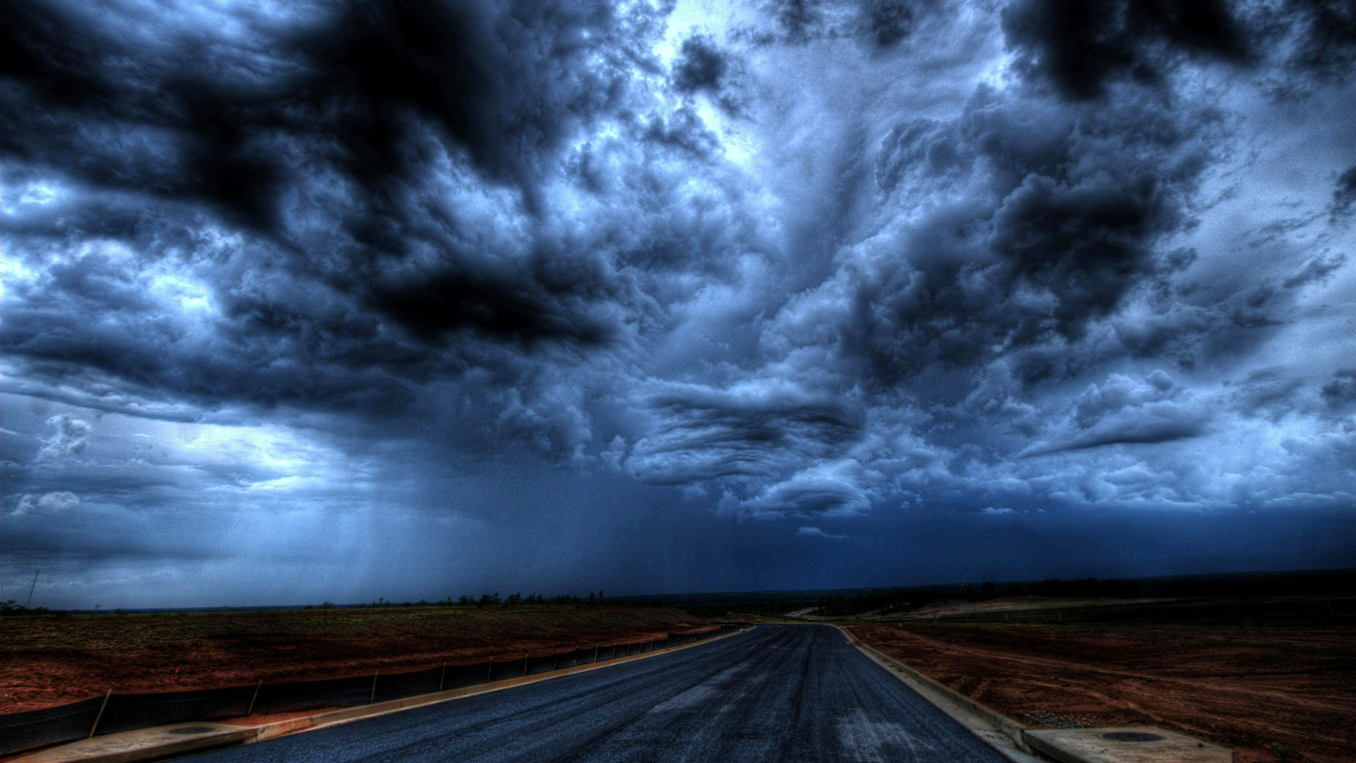 A dramatic landscape featuring a paved road stretching into the distance under a turbulent, dark, and stormy sky. Thick, swirling clouds dominate the scene, creating a sense of impending weather. The surrounding terrain is barren, with patches of dirt and grass on either side of the road, emphasizing the stark and moody atmosphere.