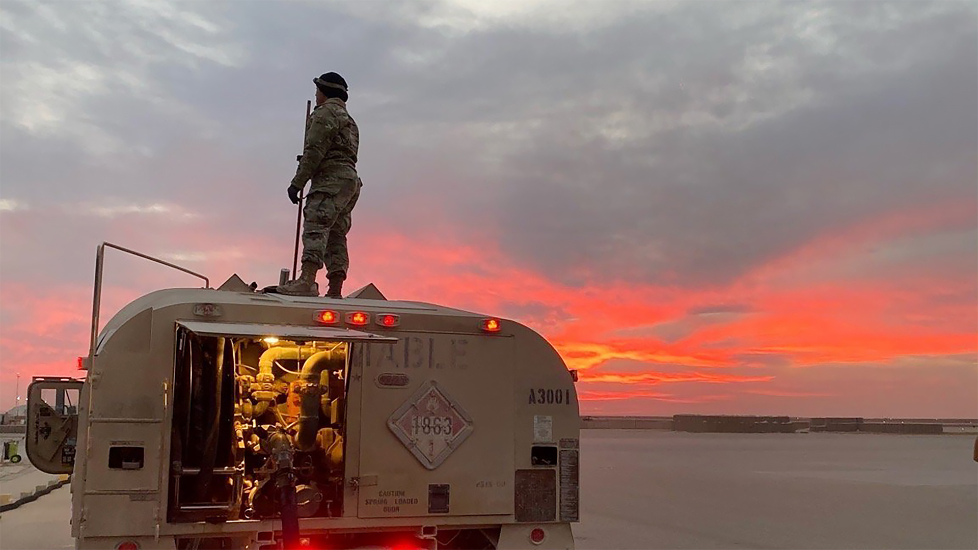 Military personnel standing on top of an armored vehicle during sunset or sunrise, with dramatic pink and orange sky in the background. The soldier is silhouetted against the colorful sky, and the vehicle appears to have its rear compartment open with lights visible inside.