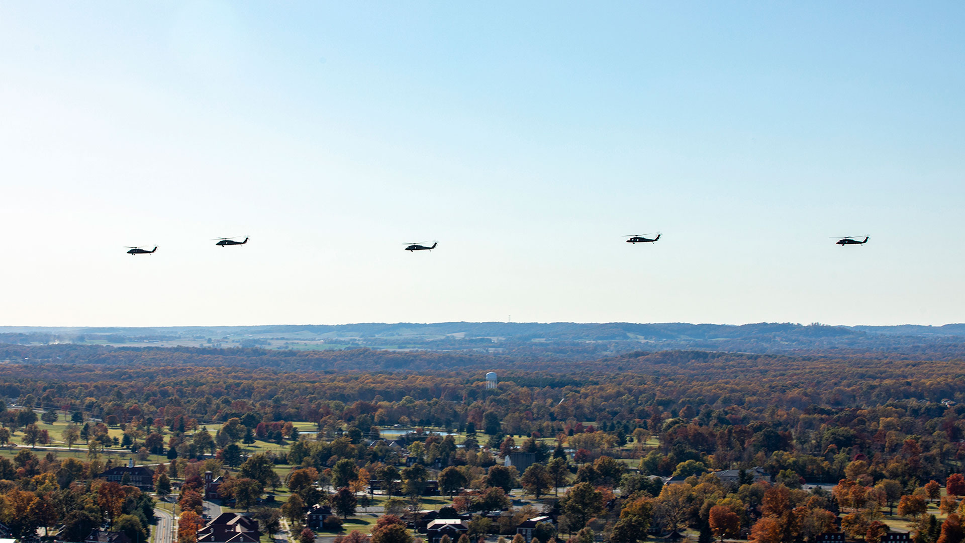 Five U.S. Army helicopters flying in formation over a landscape of autumn-colored trees and scattered buildings, with distant hills visible under a clear sky.