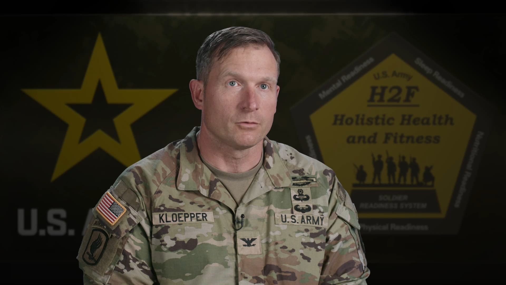 U.S. Army officer Colonel Kloepper, is positioned against a background that features the U.S. Army star logo on the left and a poster on the right displaying the words 'H2F Holistic Health and Fitness,' which is part of the Soldier Readiness System. The poster highlights aspects like mental, physical, sleep, and nutritional readiness. The officer is looking directly at the camera with a serious expression.