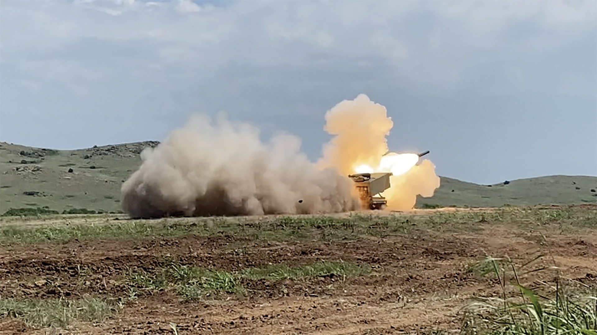  A military rocket launcher system firing in an open field, creating a large cloud of brown dust and smoke with a bright orange muzzle flash. The vehicle is positioned on sparse grassland with rolling hills in the background under an overcast sky.