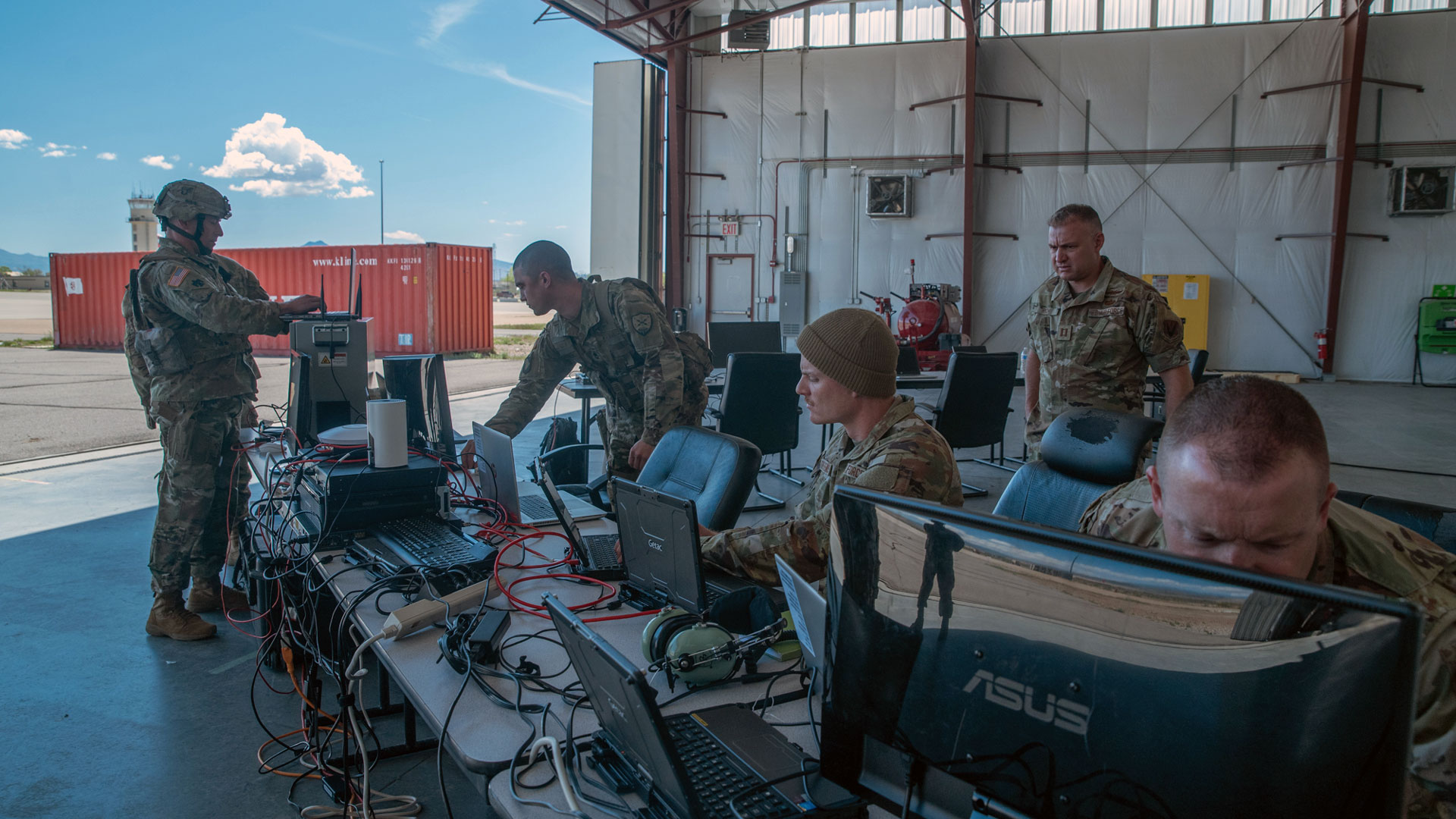 Military personnel conducting cyber operations training in a hangar. Five soldiers in combat uniforms work at computer stations with multiple monitors and equipment. Setup includes ASUS monitors and networking cables. Red shipping container and control tower visible in background.