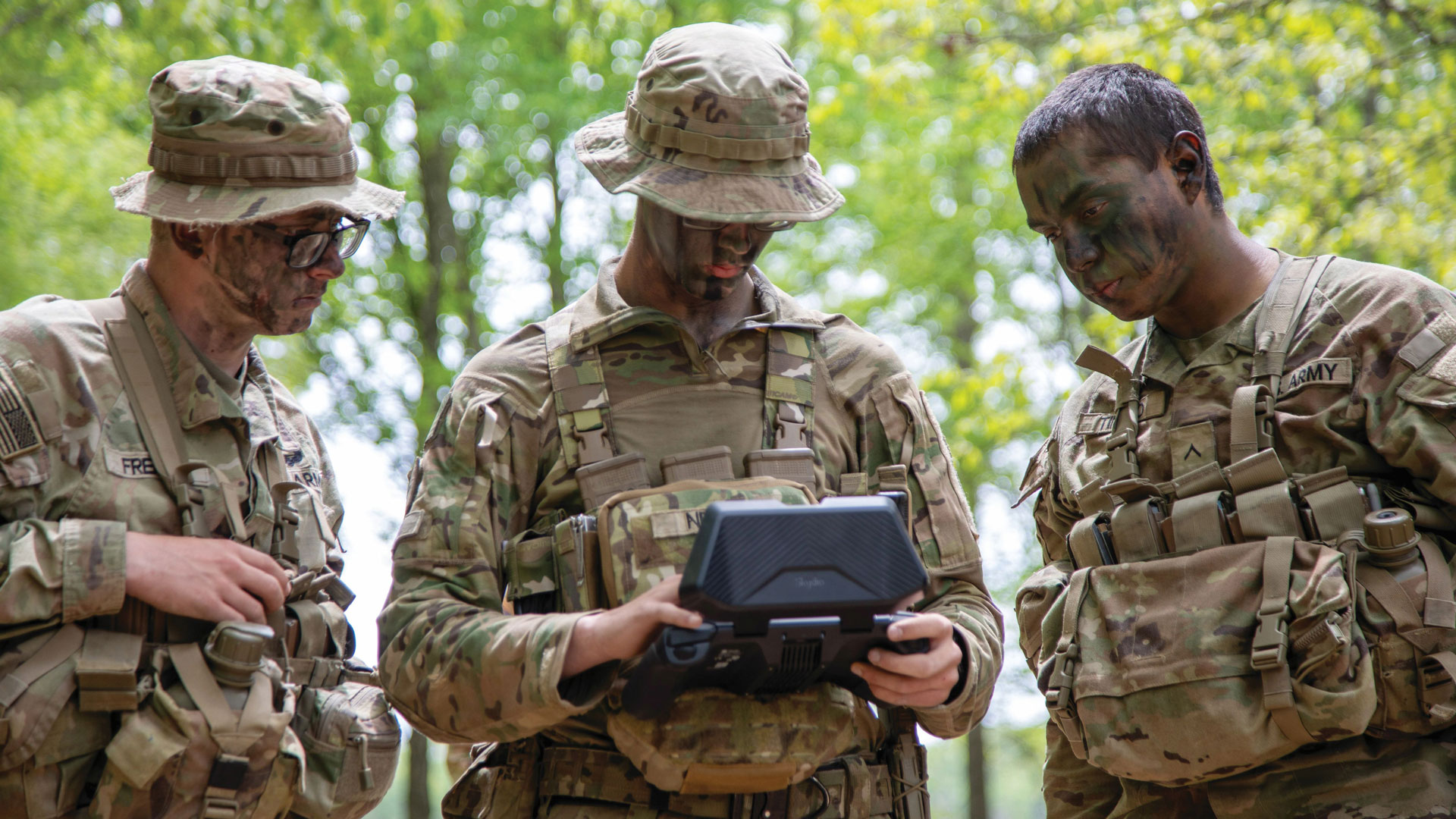 Three U.S. Army soldiers in camouflage uniforms with face paint stand in a forest, focused on a handheld controller screen, coordinating drone operations during reconnaissance training.