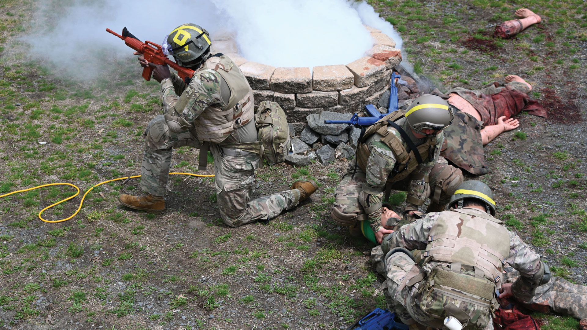 U.S. Soldiers in combat gear conduct a field medical exercise. One kneels with a training rifle and provides cover near a smoking crater, while two others give aid to a simulated casualty on the ground. In the background, a bloodied mannequin is used for realism.