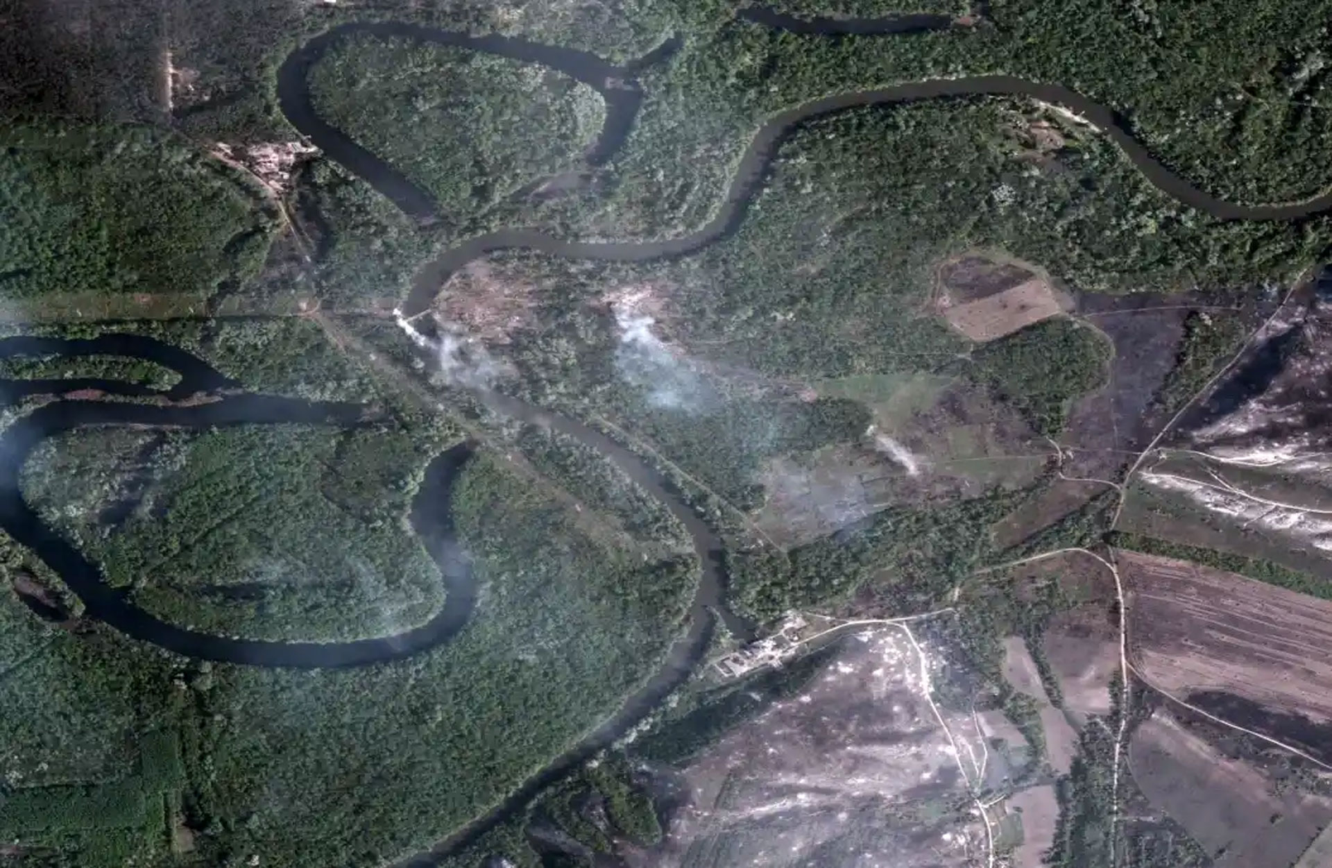Aerial view of a winding river with multiple meandering loops flowing through dense green forest, with some cleared agricultural fields and dirt roads visible in the surrounding landscape.