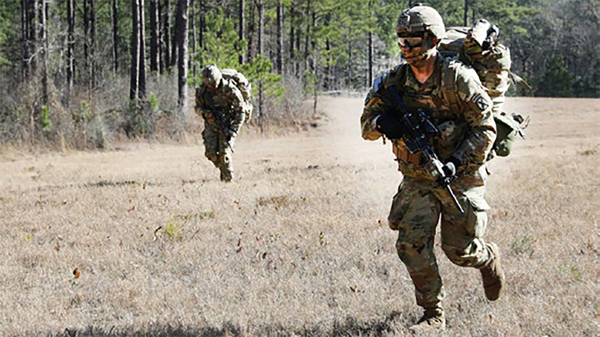 A photograph of soldiers in military combat gear running across an open field during what appears to be a training exercise. Two soldiers are visible in the foreground wearing full battle dress uniforms, helmets, and carrying weapons, with dust being kicked up as they move. Trees are visible in the background.