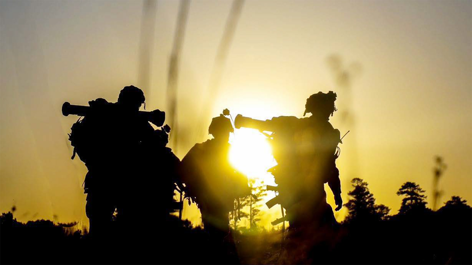 A silhouette photograph of three soldiers against a bright sunset or sunrise sky. The soldiers are carrying heavy packs and equipment, creating dramatic black silhouettes against the golden light. The image conveys the weight and burden of military gear during field operations