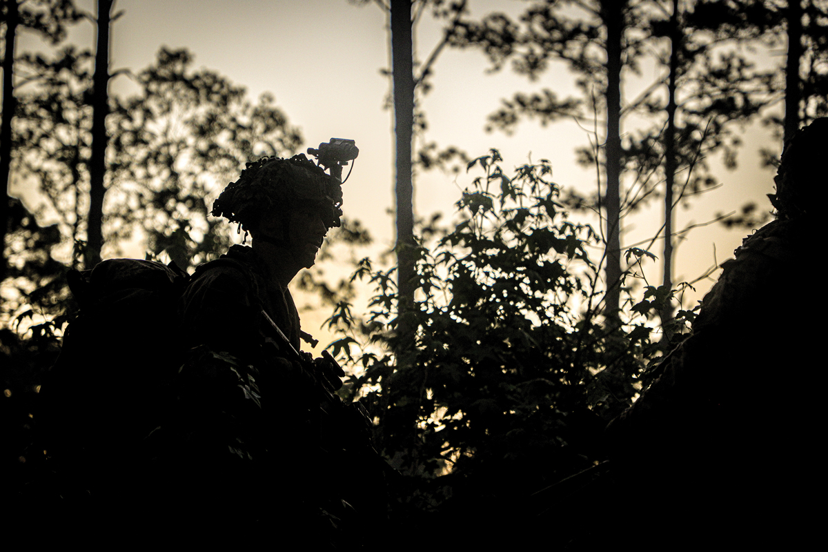 A striking image of a soldier's silhouette, equipped with night vision gear, standing amidst dense foliage in a forest at dusk. The backdrop of the setting sun and tall trees highlights the soldier's vigilance and readiness.