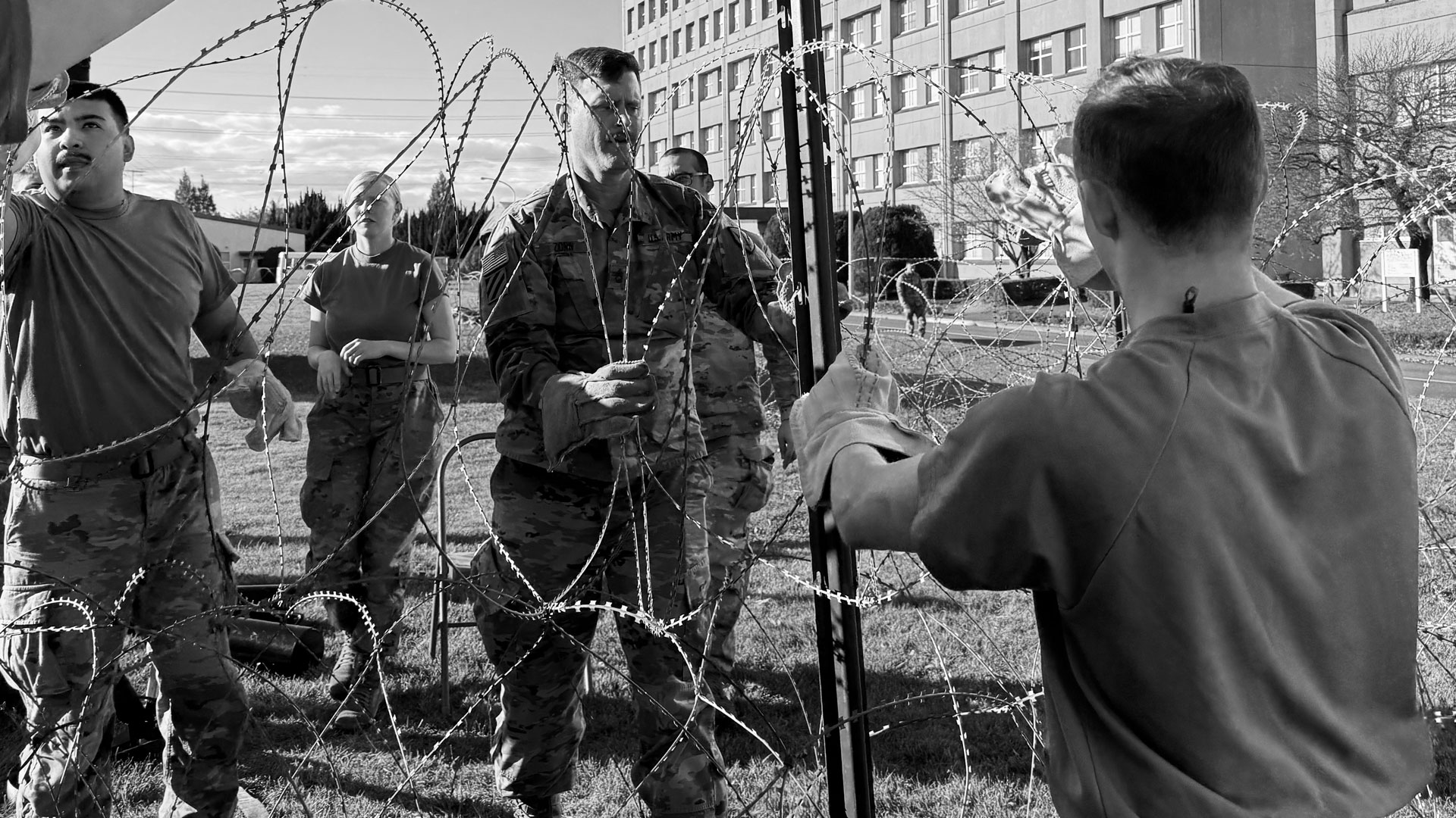 Black and white photo of U.S. military personnel in uniform installing concertina wire outside a building. One soldier is holding the wire while others assist in positioning it. A large multi-story building and clear sky are visible in the background.