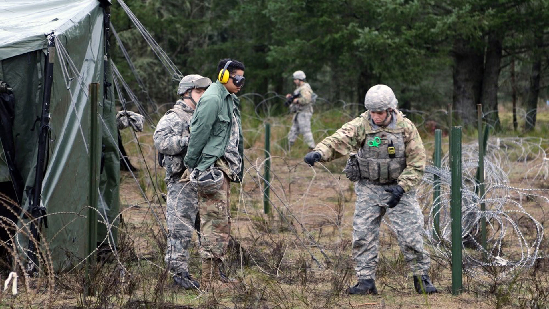 A group of soldiers in military uniforms conducting detainee operations training in a wooded area. One soldier is escorting a detainee wearing earmuffs near a tent and barbed wire fencing, while another soldier gestures and provides instructions. Additional soldiers are visible in the background near the perimeter. The scene emphasizes a controlled training environment.