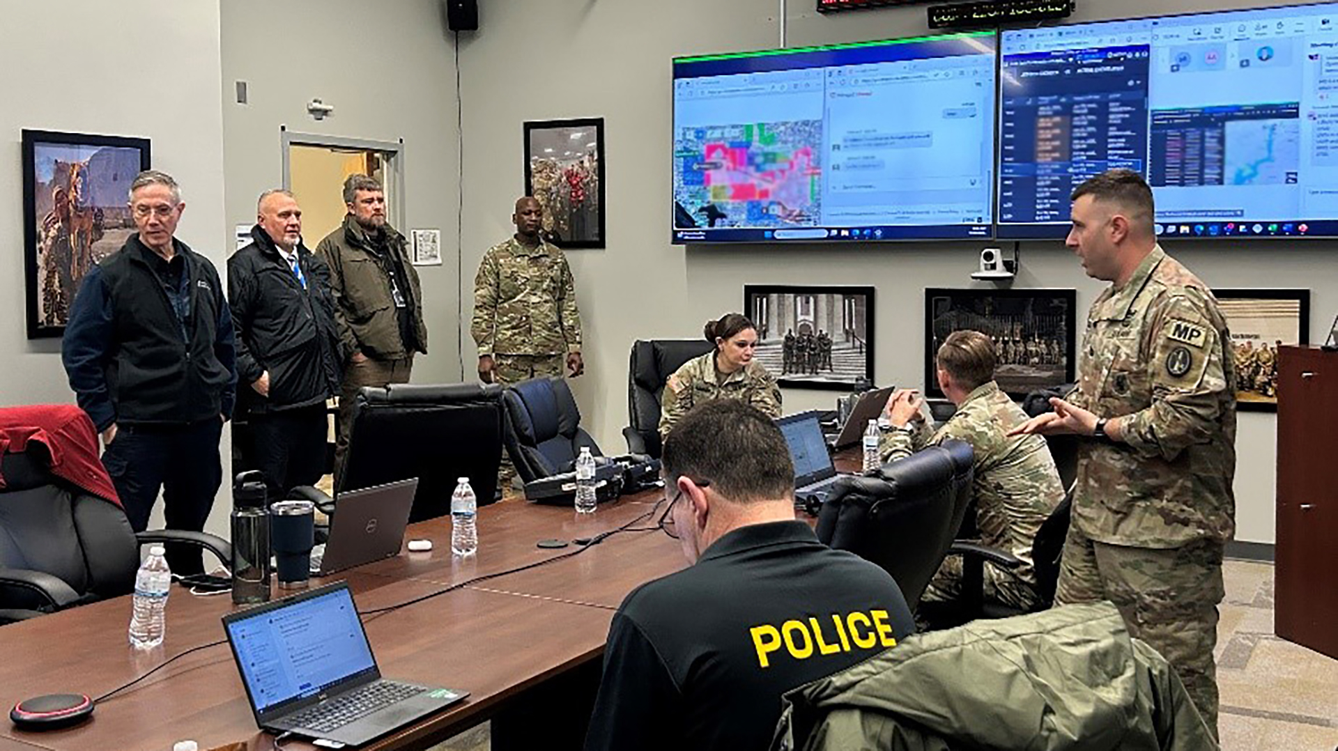 Military personnel and police officers in a command center or briefing room with multiple computer screens, digital displays, and conference table during an operational meeting