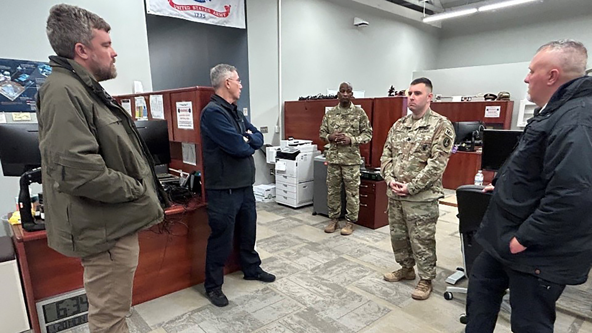 Military personnel in multicam uniforms meeting with civilian officials in business attire inside an office space with cubicles and a United States banner visible in the background