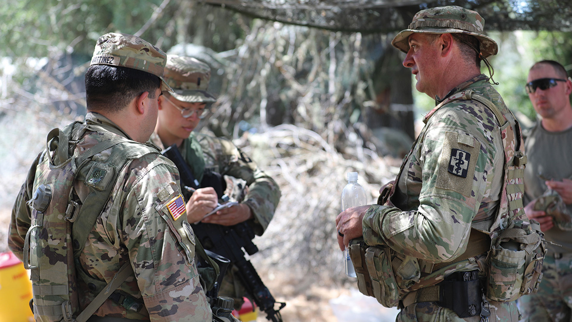 Three soldiers in camouflage uniforms standing outdoors, engaged in conversation under partial shade.