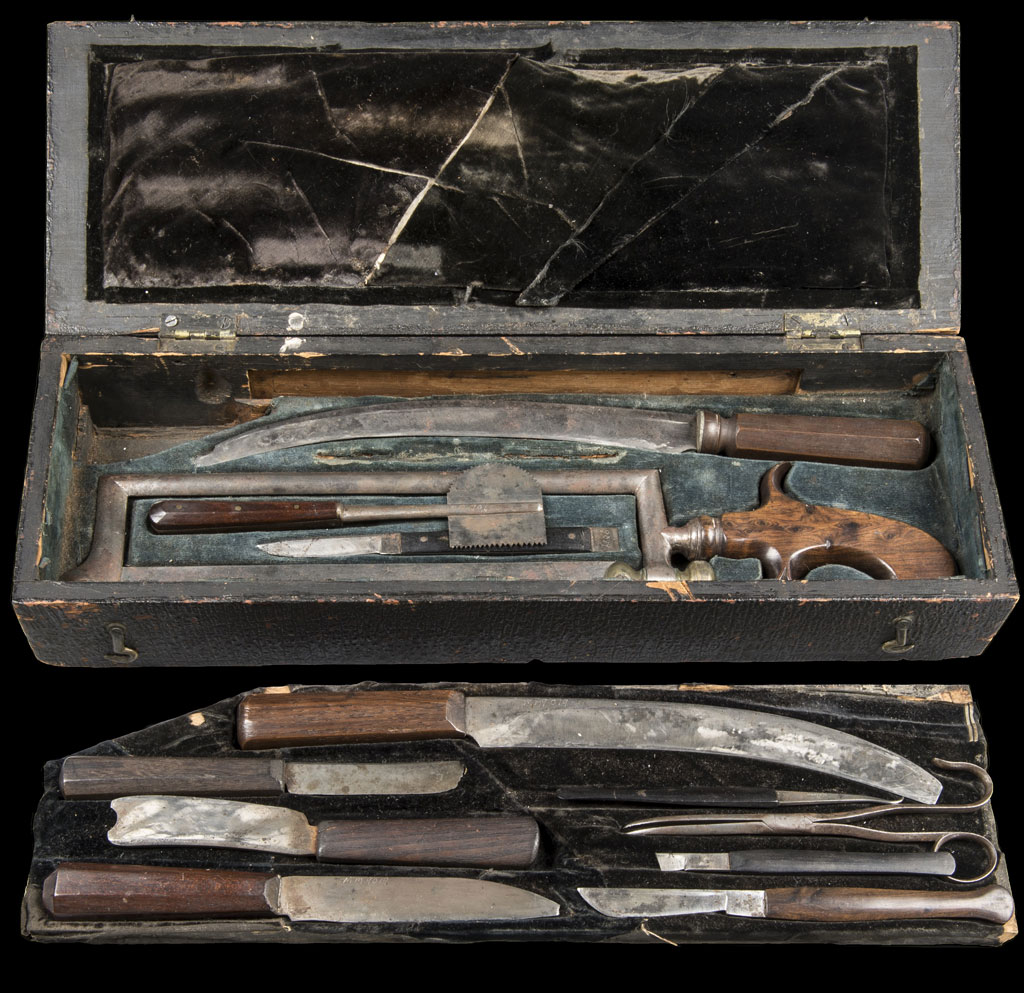 A historic surgical kit displayed in an open black box, containing various vintage medical instruments including knives, saws, and a small pistol. The tools have worn wooden handles and are organized in velvet-lined compartments.