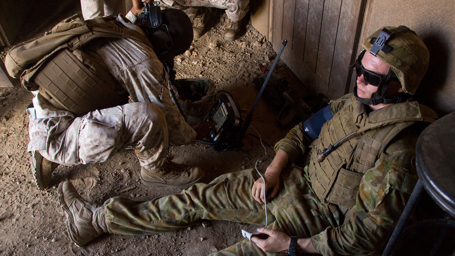 A military medic kneels beside an injured soldier, operating a medical device in a dimly lit, dusty environment. The injured soldier sits against a wall, connected to medical equipment, while wearing camouflage and protective gear.