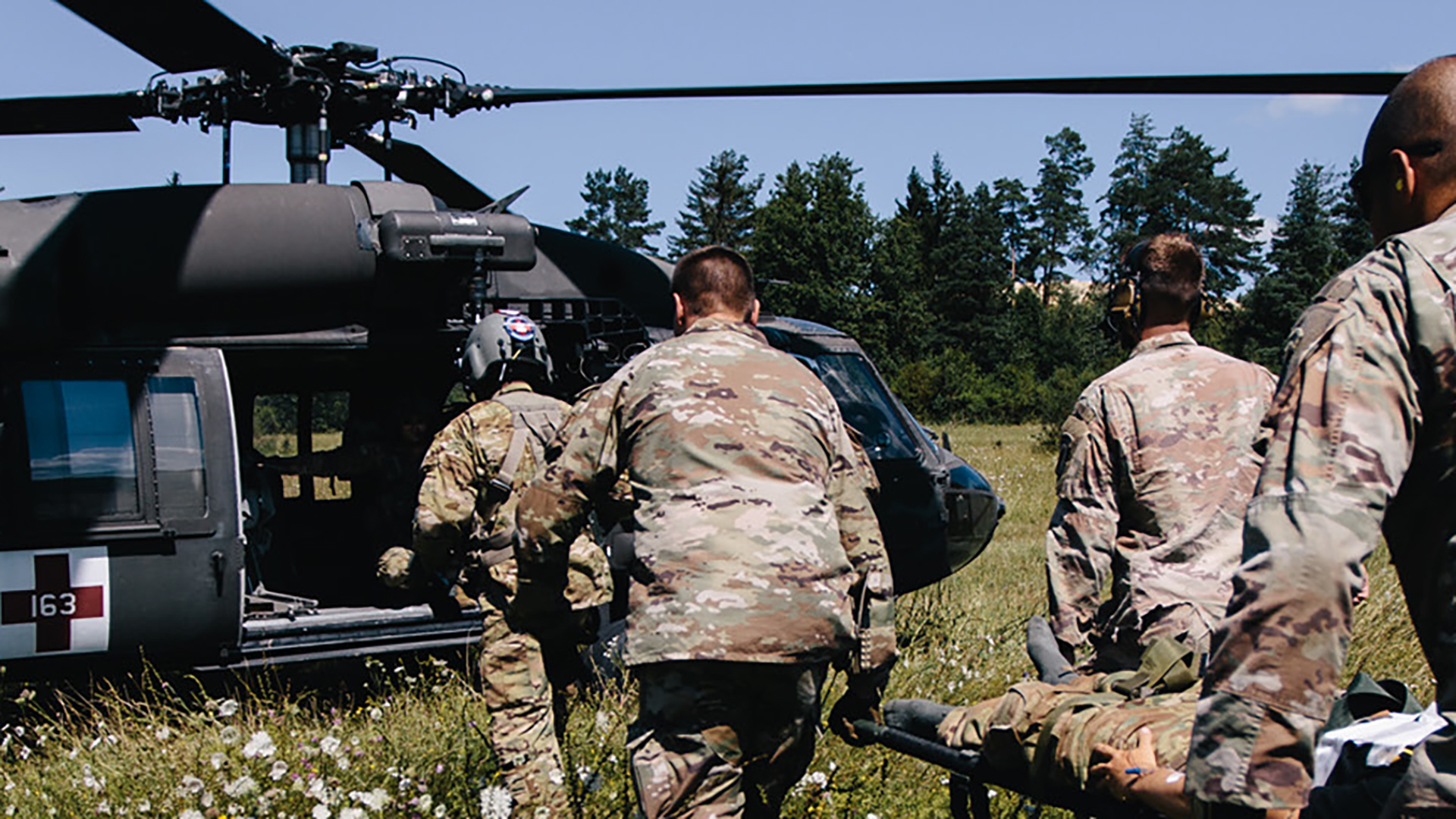 Military personnel in desert camouflage uniforms running toward a military helicopter (appears to be a UH-60 Black Hawk) that has landed in a grassy field. The soldiers are carrying equipment and appear to be conducting a medical evacuation or transport exercise, with pine trees visible in the background.