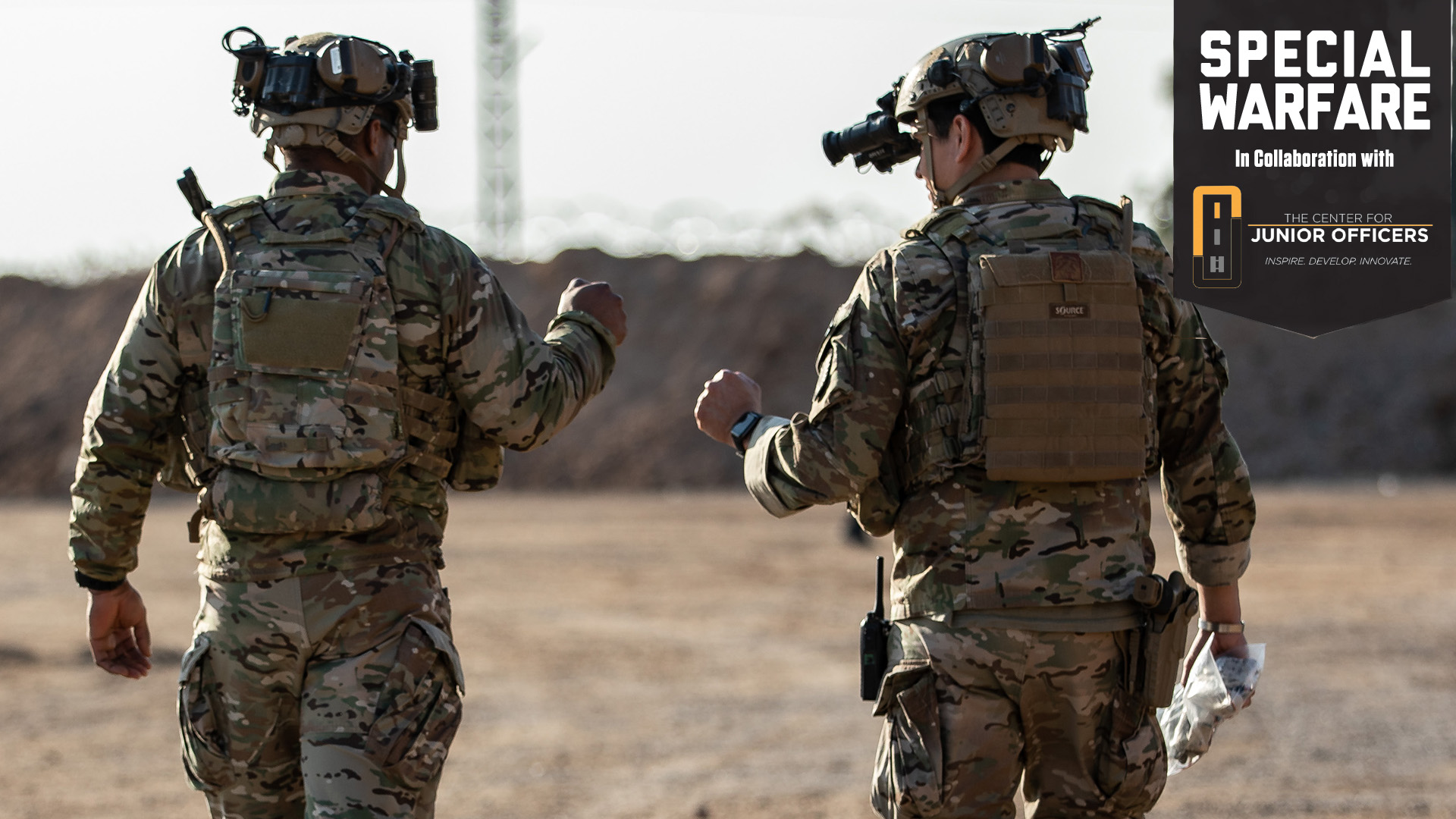 Two soldiers in multicam camouflage uniforms walking away from the camera across a dusty, arid landscape. Both are wearing tactical helmets equipped with night vision goggles and other electronic devices, along with full combat gear including body armor and tactical vests. The soldier on the right appears to be holding or carrying something in their hands. The background shows a blurred desert or training environment with some structures visible in the distance.