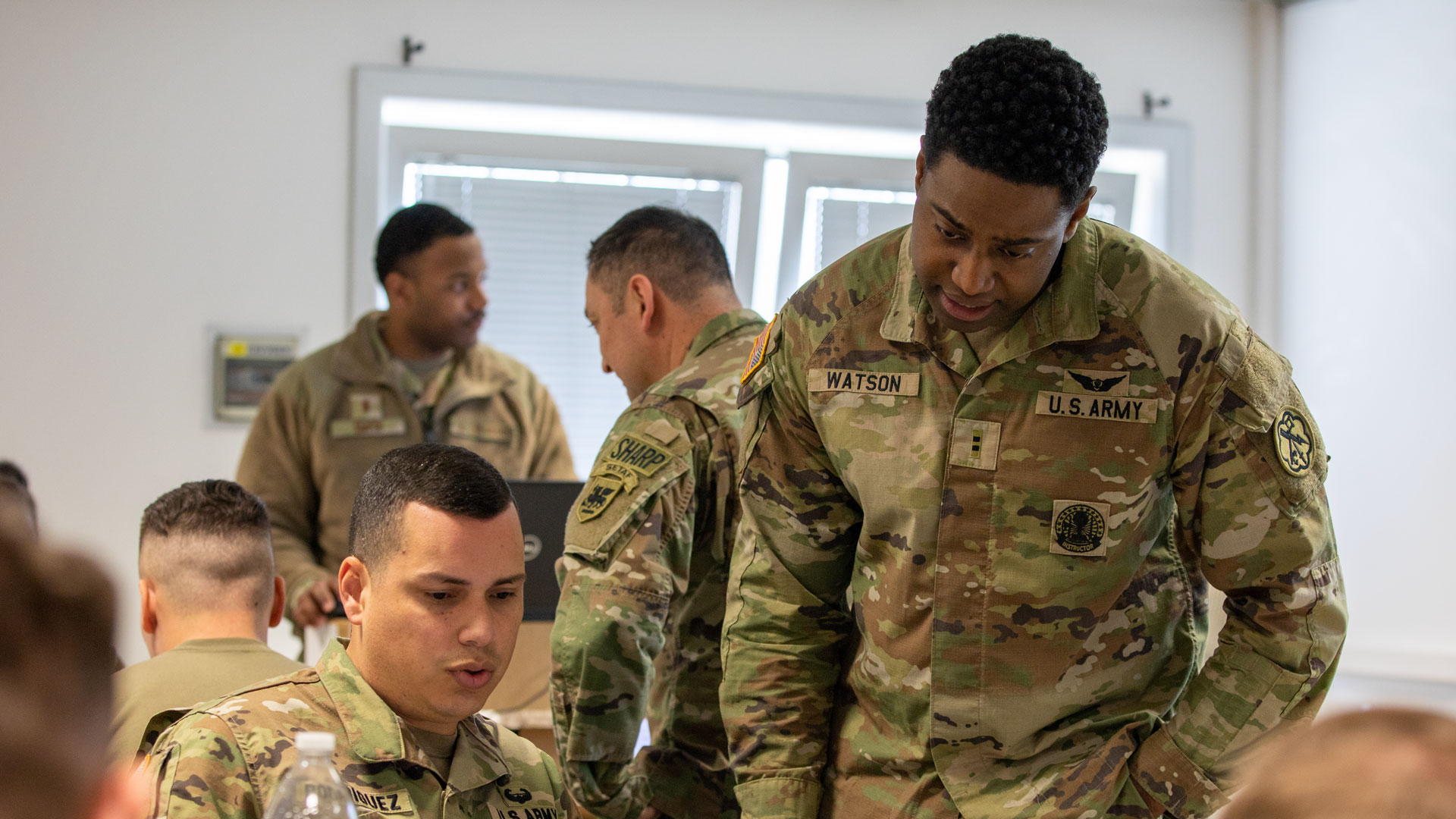 U.S. Army Warrant Officer in uniform leans over to discuss with a fellow soldier seated at a table, as other service members work in the background during a joint task force exercise in a classroom setting.