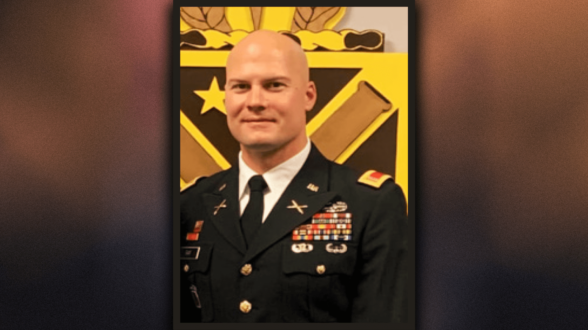 A military officer dressed in a formal uniform adorned with multiple ribbons, medals, and insignia. The background features a yellow and black emblem with crossed cannons and a star.
