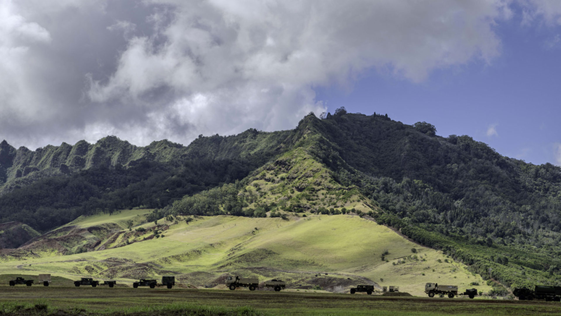 mountainous landscape with green foliage and a cloudy sky. A line of military vehicles is spread across a grassy field in the foreground