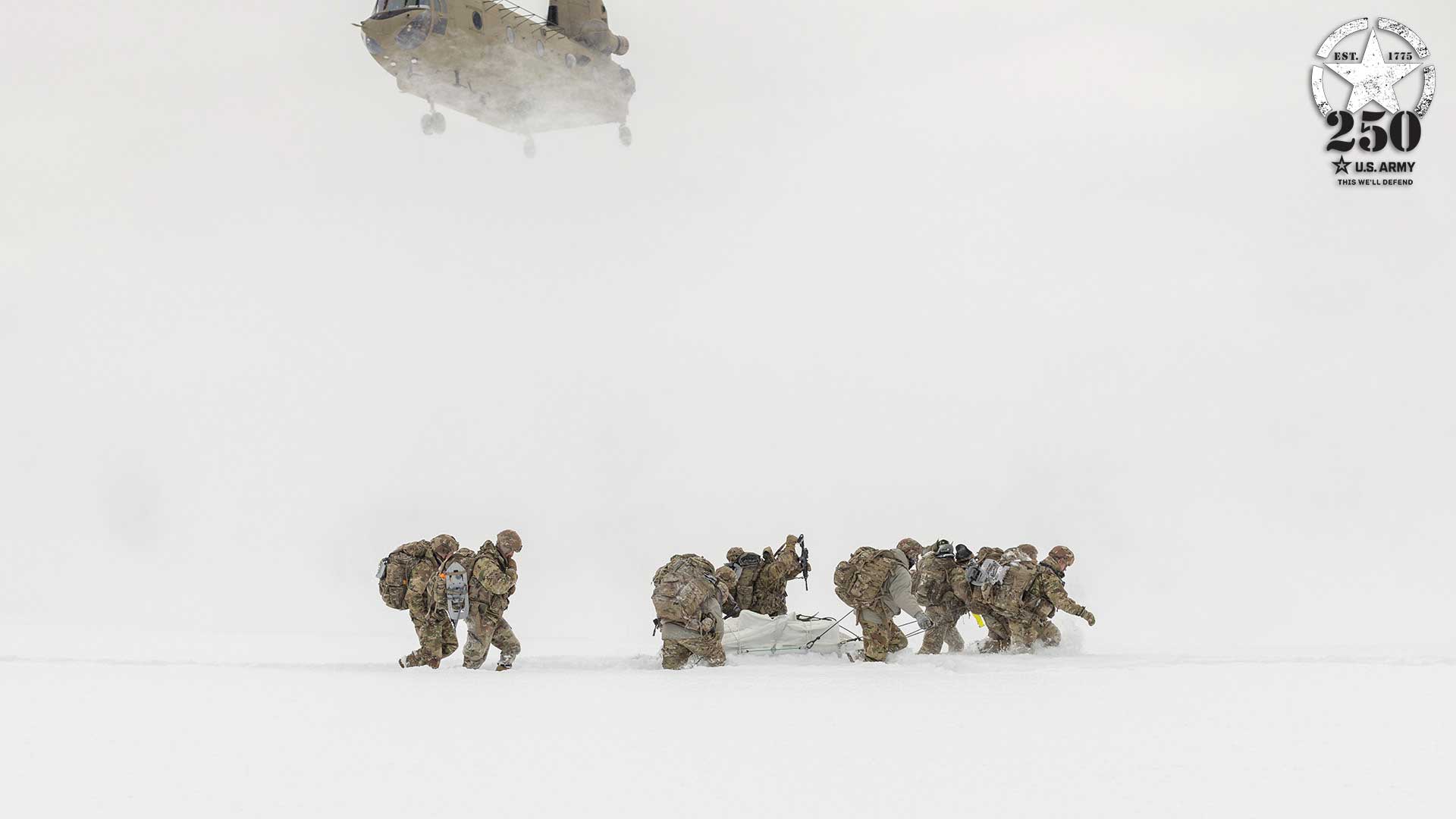 U.S. Army soldiers in winter combat gear conducting medical evacuation training in snow-covered terrain. Group of soldiers pulling casualty on litter while CH-47 Chinook helicopter hovers above creating snow cloud. U.S. Army 250th anniversary emblem visible in top right corner.